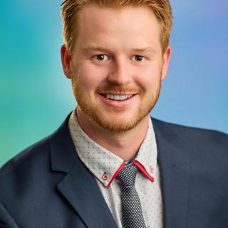 Mitch Turko wearing a suit and tie, smiling at the camera with a blue and green background.