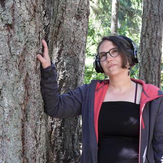 Sharon leans against a tree wearing headphones