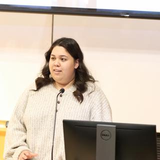 A student wearing a white sweater stands in front of a microphone giving their presentation.