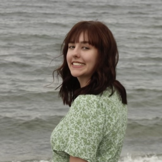 Paige Pierce standing in front of the ocean, wearing a green dress and turning back to smile at the camera