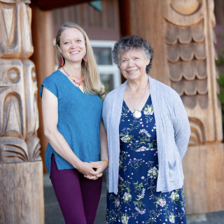 VIU Psychedelic-assisted Therapy Program Chair Shannon Dames with VIU Elder-in-Residence C-tasi:a Geraldine Manson standing outside in front of totem poles and smiling at the camera