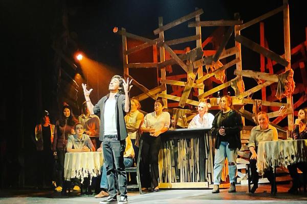 An actor at centre stage has their hands thrown up in the air while they look off in the distance. Numerous cast mates stand behind them in various poses.