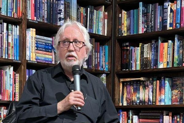 Perry Bulwer speaks into a microphone with a shelf of books behind him