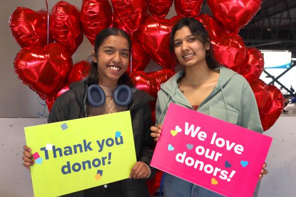 Two students hold up signs saying thank you donor and we love our donors and stand in front of red heart balloons