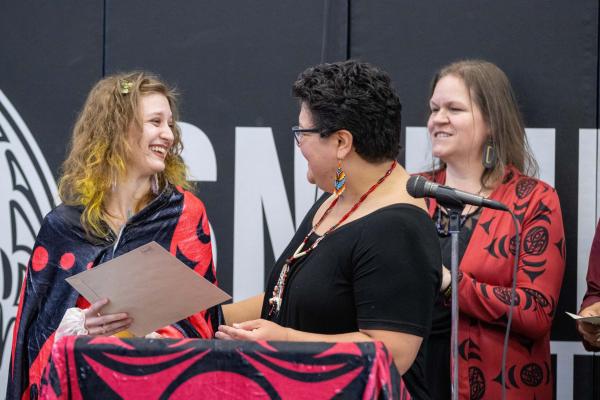 A student receives a folder from a woman, with another woman watching in the background