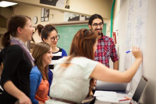 A group of students gathers around a student writing on a chalkboard