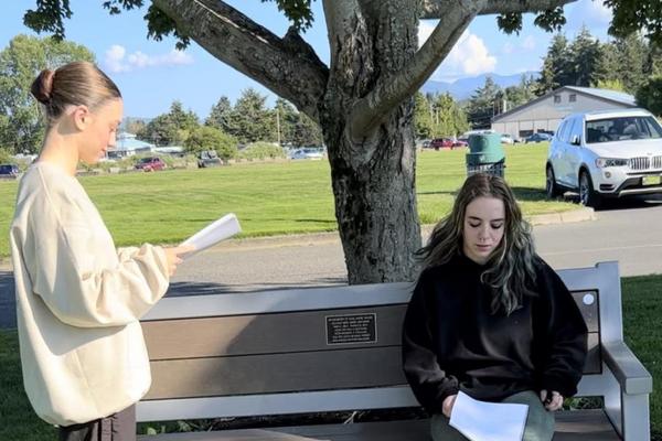 Two actors rehearse with scripts in their hand near a bench in a park.