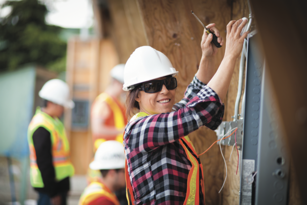 Female electrician wearing. white hard hat a safety vest and sunglasses working on the side of a building and smiling at the camera.