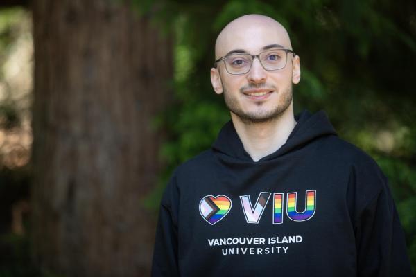 VIU grad and current employee Rayan Zeineddine wearing a VIU hoodie and glasses standing next to a tree outside and smiling at the camera