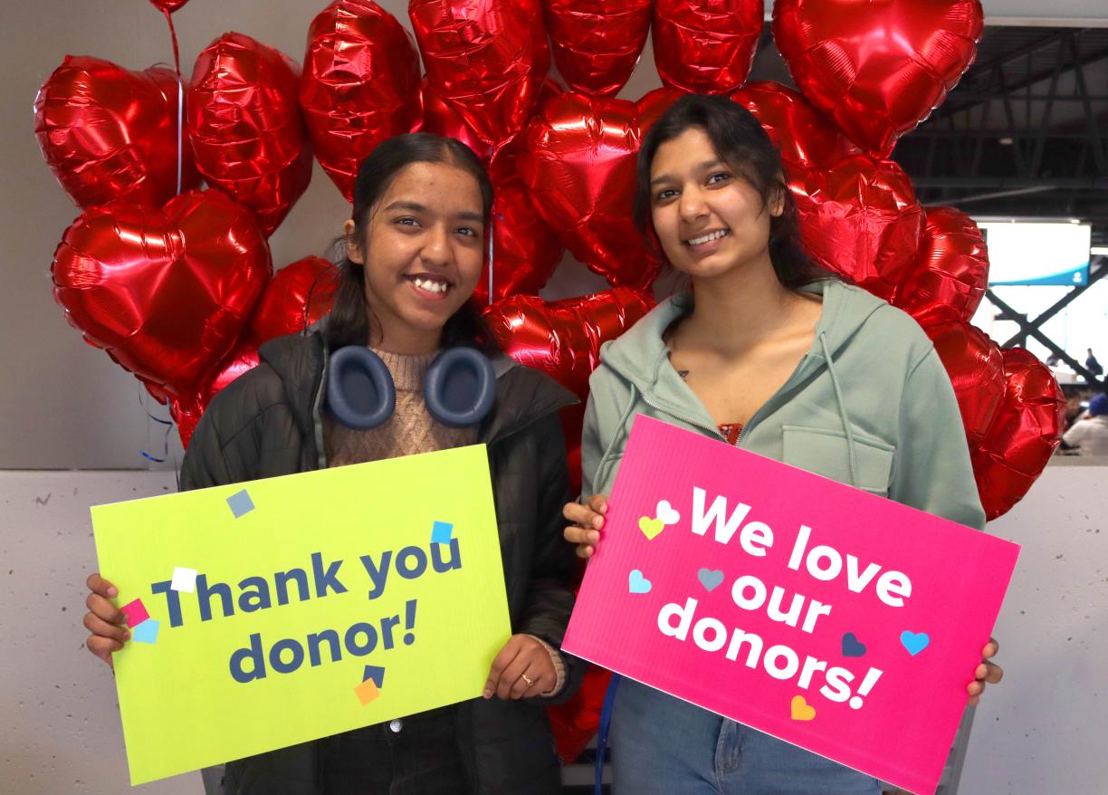 Two students hold up signs saying thank you donor and we love our donors and stand in front of red heart balloons