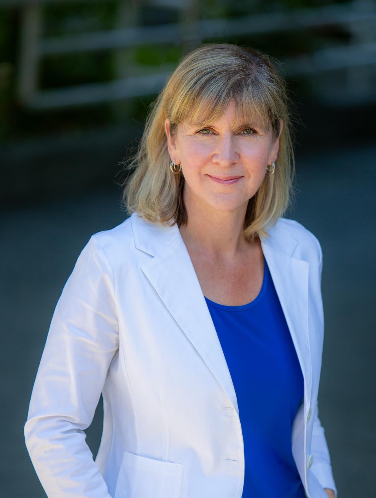 Kristie Dewald with a blurred background, wearing a white blazer