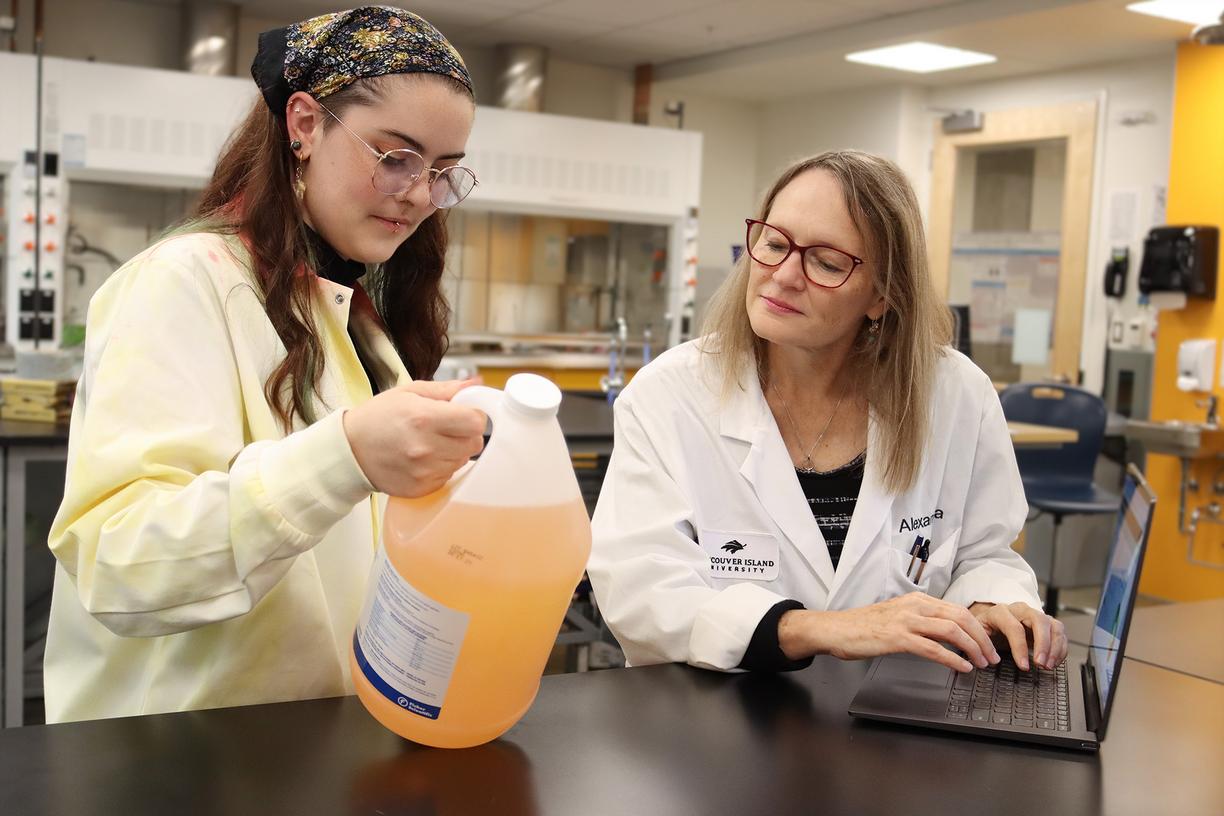 Lily Eggert and Dr. Alexandra Weissfloch look at a bottle of cleaning product, which is an amber colour, while in a VIU chemistry lab. Both wear lab coats.