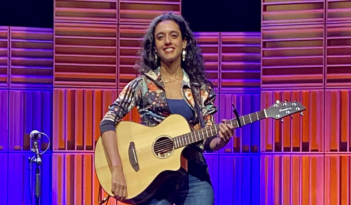 Sonnet L’Abbé sits on a stool on the Port Theatre's stage in front of a multi-coloured pink, blue and purple lighted background.