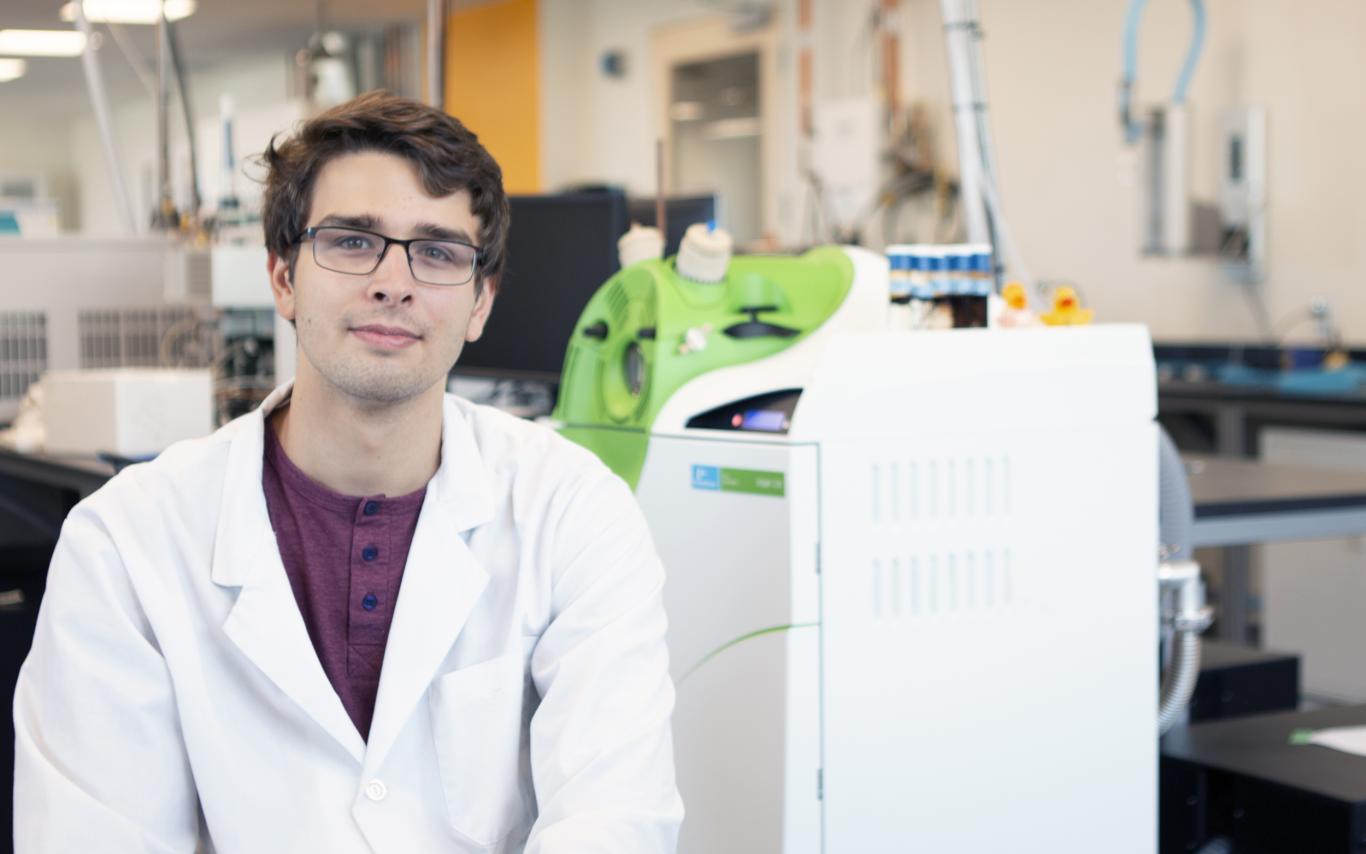 Joseph Monaghan sits in a VIU Chemistry lab, wearing a white lab coat.
